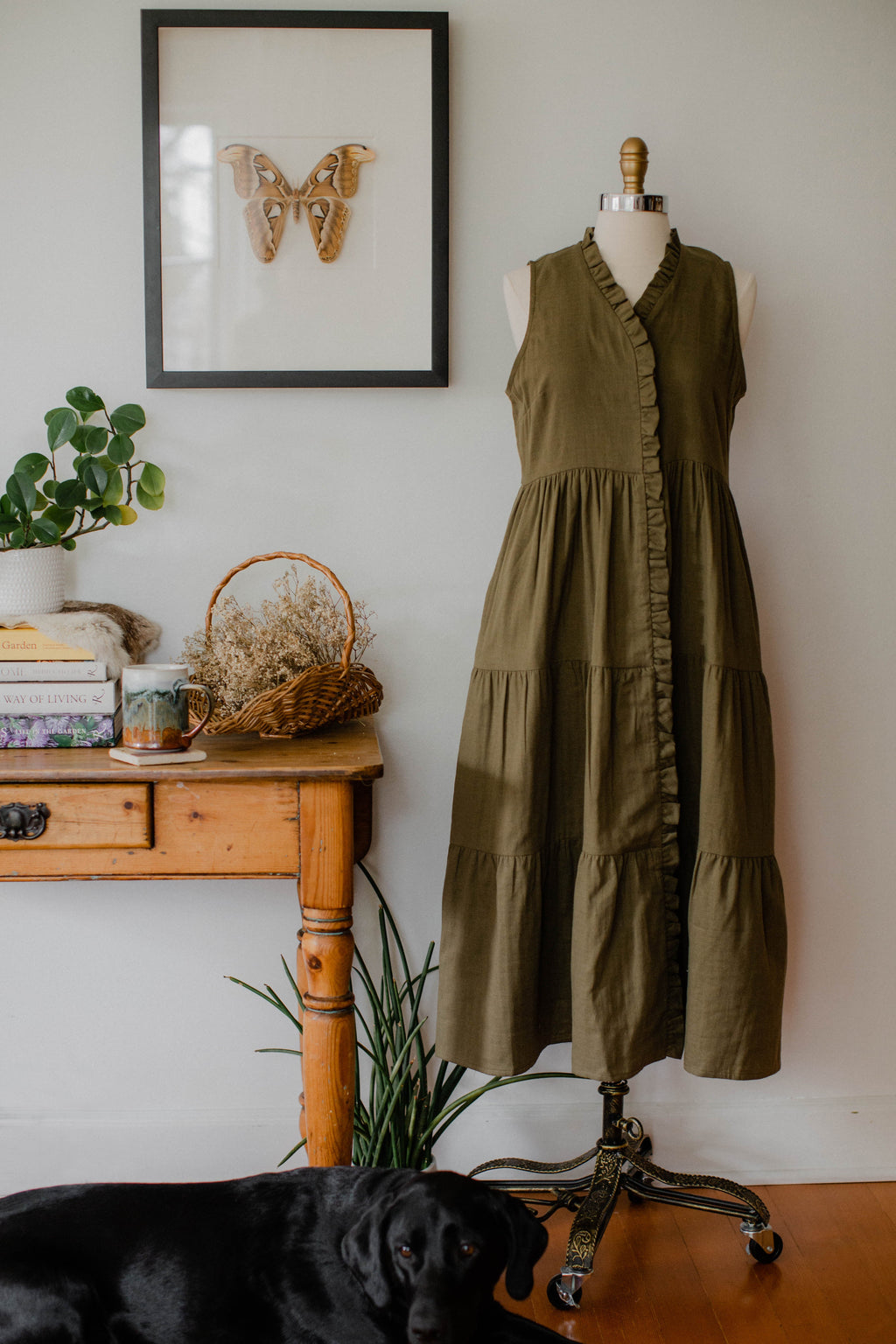 The House Dress displayed on a dress form in an earthy green color in a cozy, decorative room setting