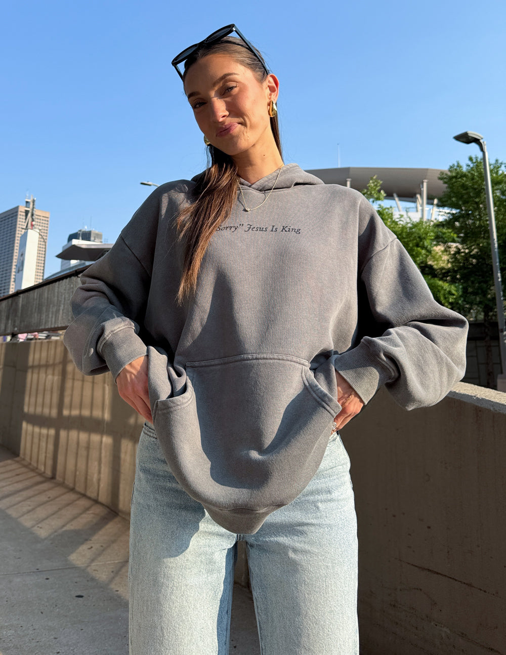 Woman wearing gray Not Sorry Jesus Is King Unisex Hoodie standing outdoors with sunglasses on her head