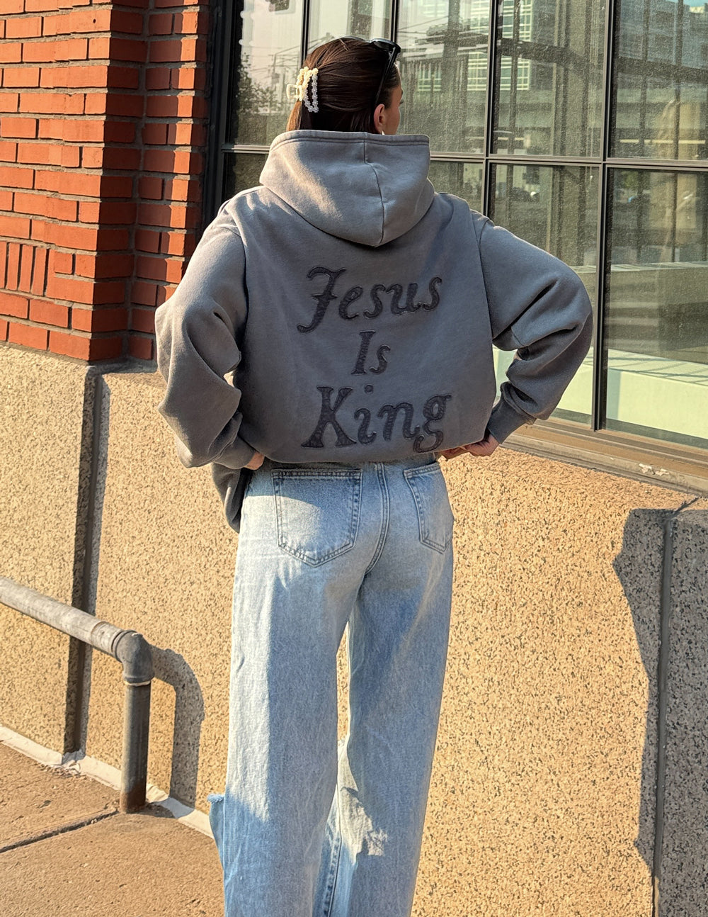 Person wearing a gray "Not Sorry Jesus Is King Unisex Hoodie" with bold text on the back standing outdoors near a building