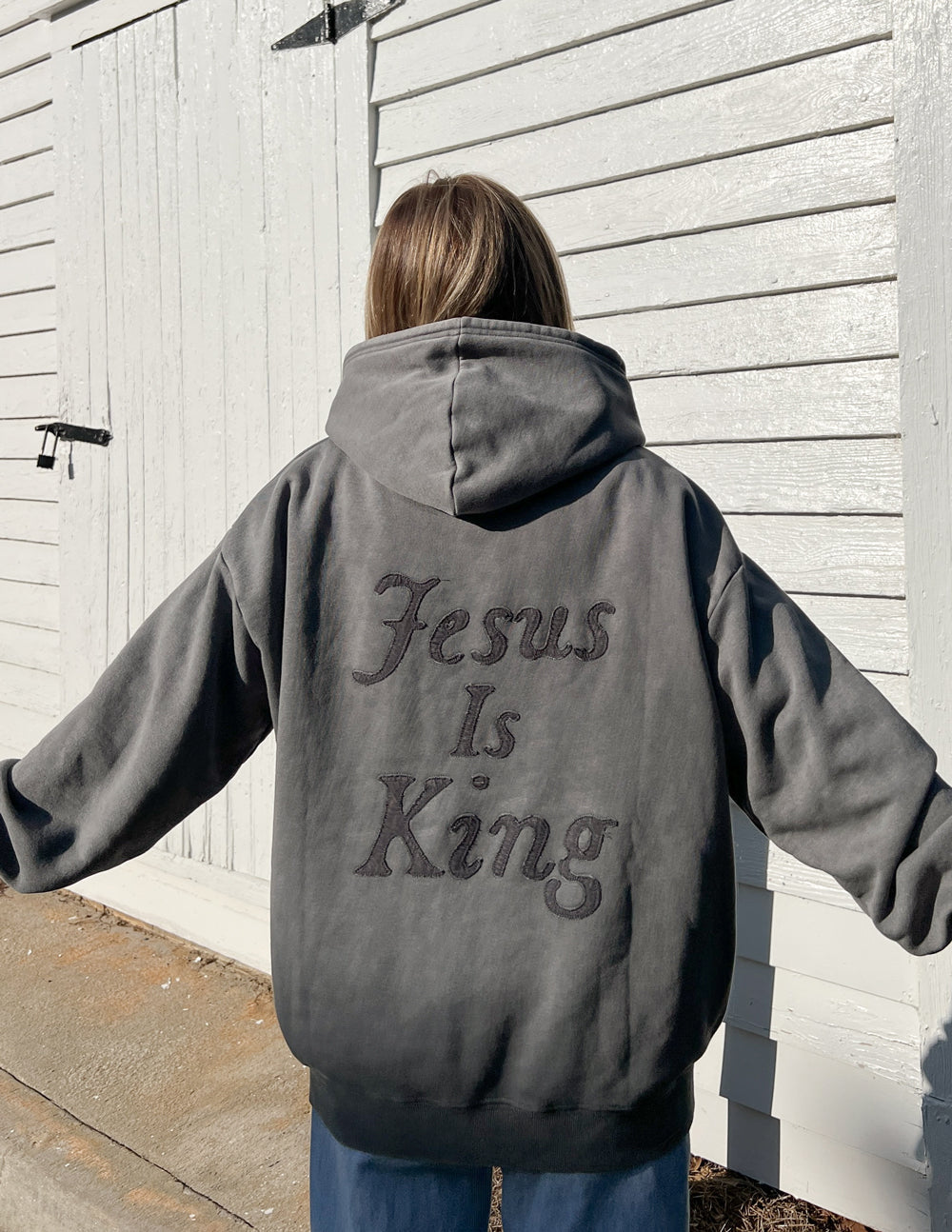 Person wearing Not Sorry Jesus Is King Unisex Hoodie in gray with large embroidered text on the back outdoors