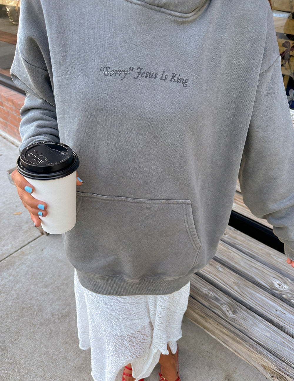 Person wearing gray Not Sorry Jesus Is King Unisex Hoodie holding a coffee cup outdoors near wooden bench.