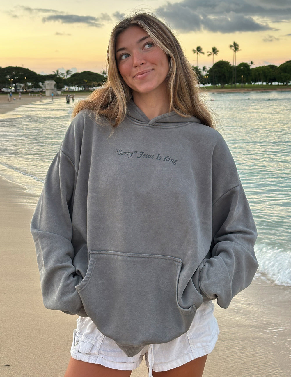 Young woman wearing a gray Not Sorry Jesus Is King Unisex Hoodie standing on a beach at sunset
