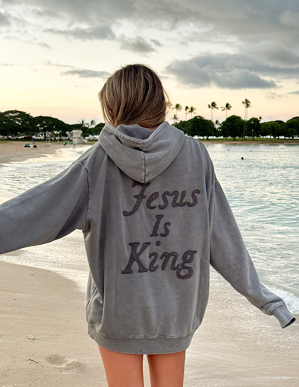 Person wearing gray Not Sorry Jesus Is King Unisex Hoodie with text on the back standing on a beach at sunset.