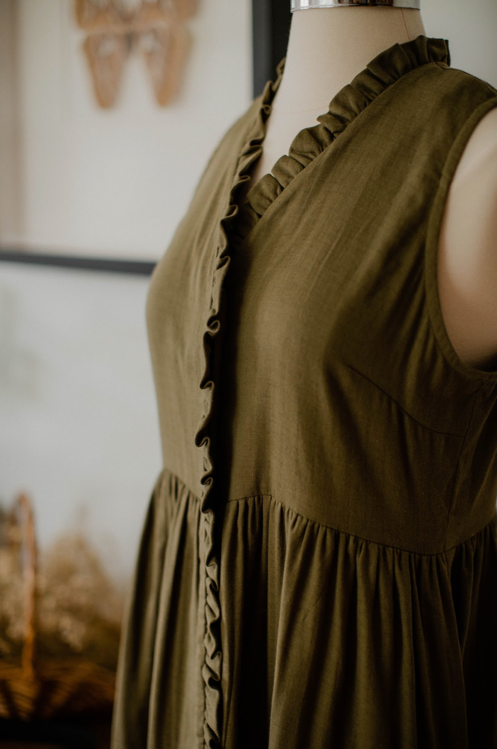 Close-up of an olive green sleeveless dress with ruffled button trim on a mannequin titled The House Dress