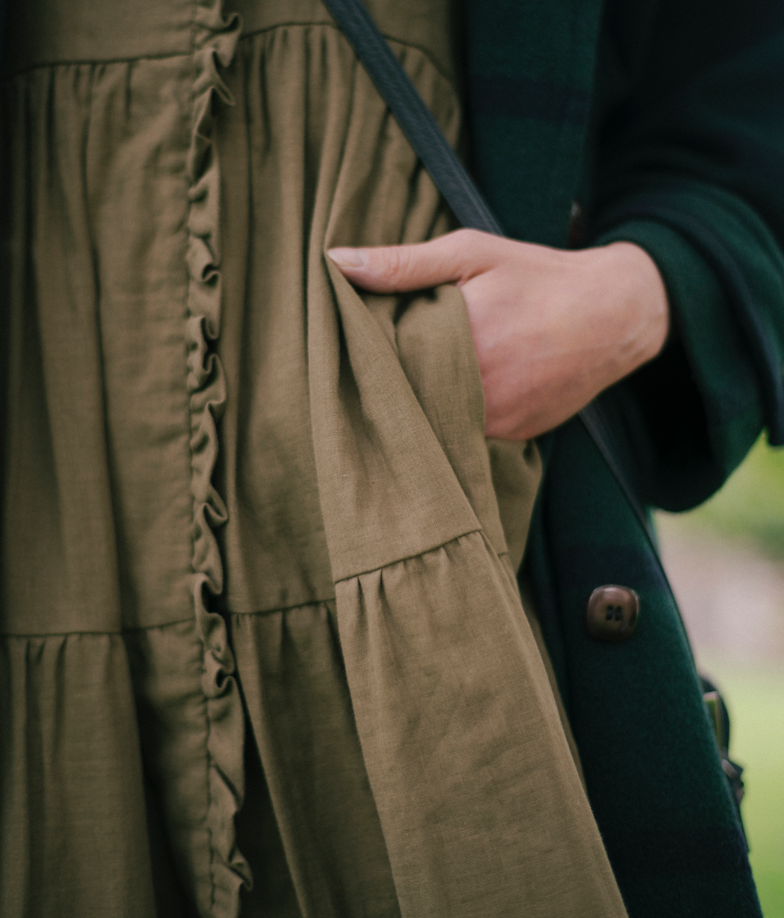 Close-up of a hand in the pocket of a brown gathered fabric dress, The House Dress, worn with a dark coat.