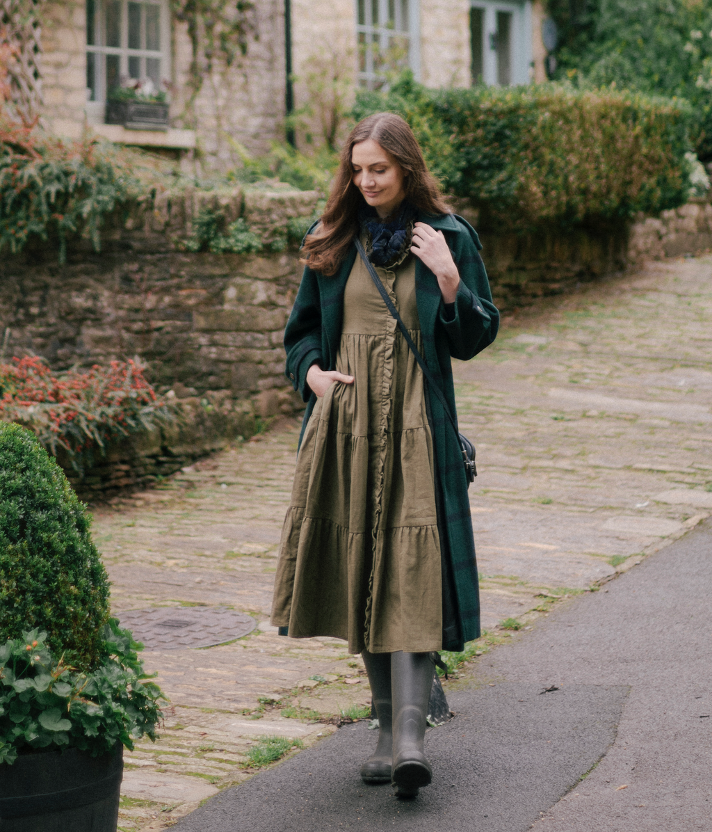 Woman wearing The House Dress in olive green with pockets, walking outdoors on a cobblestone path in a dark coat.