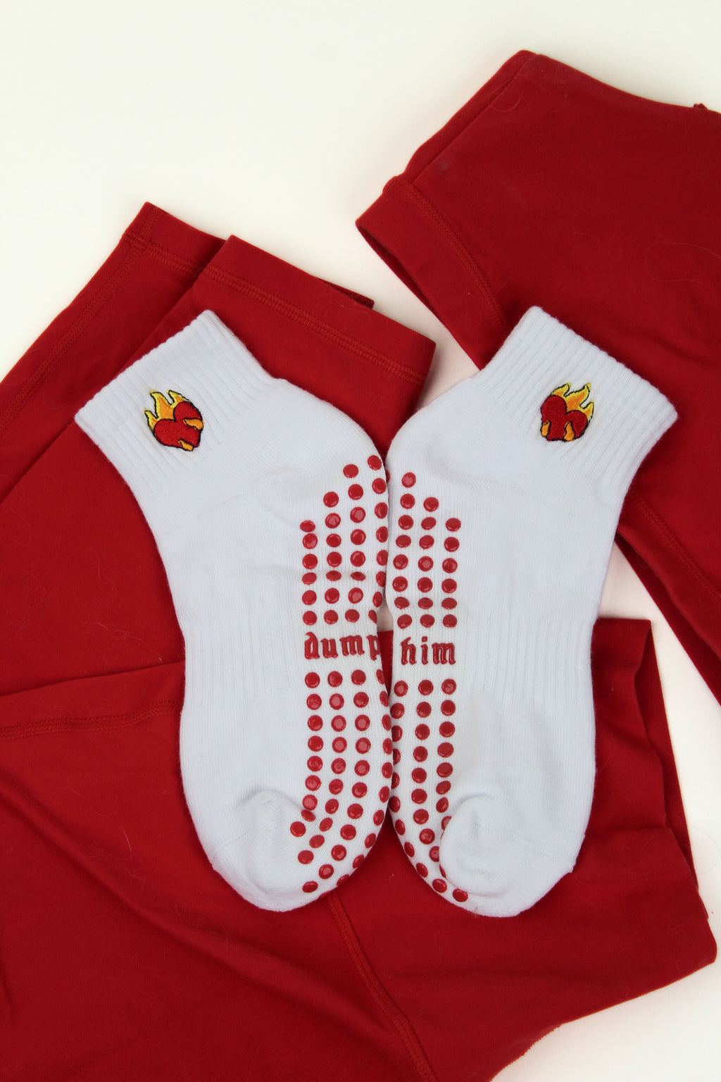 White Dump Him Grip Socks with red heart flame design and red anti-slip dots on soles, laid on red fabric background